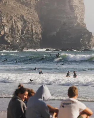 a group of people sitting on the beach watching surfers at The Suites by The Lighthouse Arrifana in Aljezur
