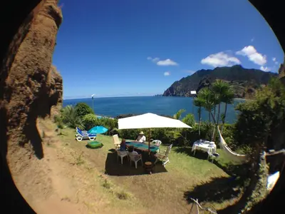 a person sitting at a table with an umbrella next to the ocean at Maresia Gardens in Porto da Cruz