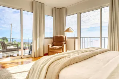 a bedroom with a bed and a chair and large windows at Ponta do Sol Sea View, a Home in Madeira in Ponta do Sol
