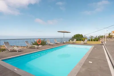 a swimming pool with chairs and an umbrella and the ocean at Villa Panoramica II in Arco da Calheta