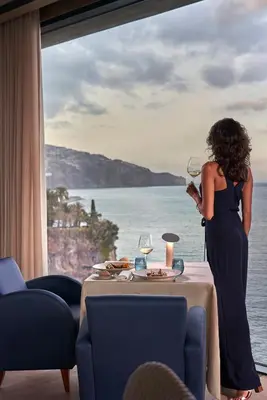 a woman standing in front of a window drinking wine at Les Suites at The Cliff Bay - PortoBay in Funchal