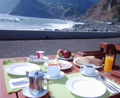 a table with plates of food on a table with the beach at Hotel Vila Bela in Porto da Cruz
