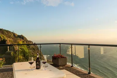 a table with two glasses of wine on a balcony at OurMadeira - Sunset Cliff Villas in Fajã da Ovelha